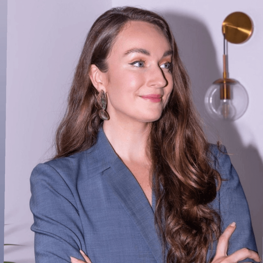 Woman in a blue blazer standing with arms crossed against a light-colored wall.