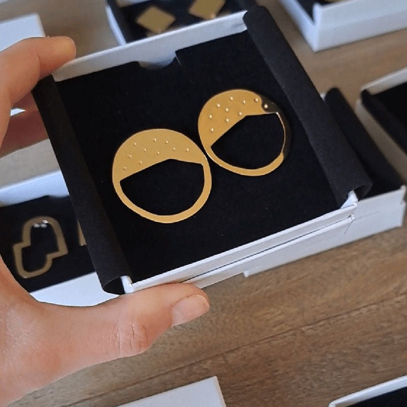 Gold circular Galaxy earrings designed by Bold Lines Jewellery are shown in its packaging, a white box, and a black sleeve.