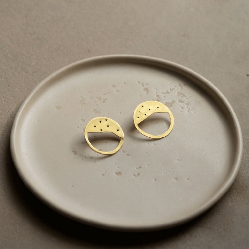 Gold circular Galaxy earrings designed by Bold Lines Jewellery are shown on a handmade beige ceramic plate.