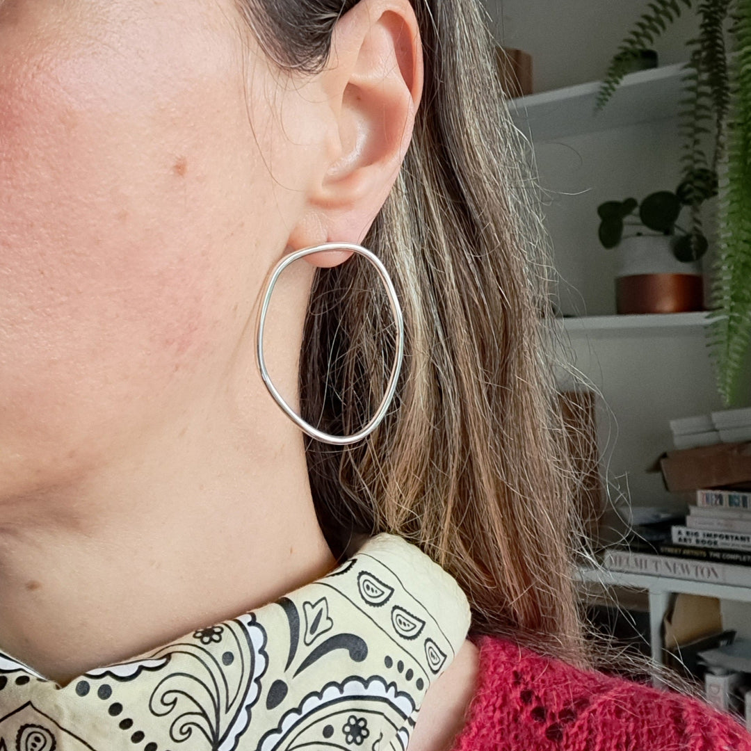 Close-up of a person wearing wavy sterling silver hoop earrings with a blurred indoor background