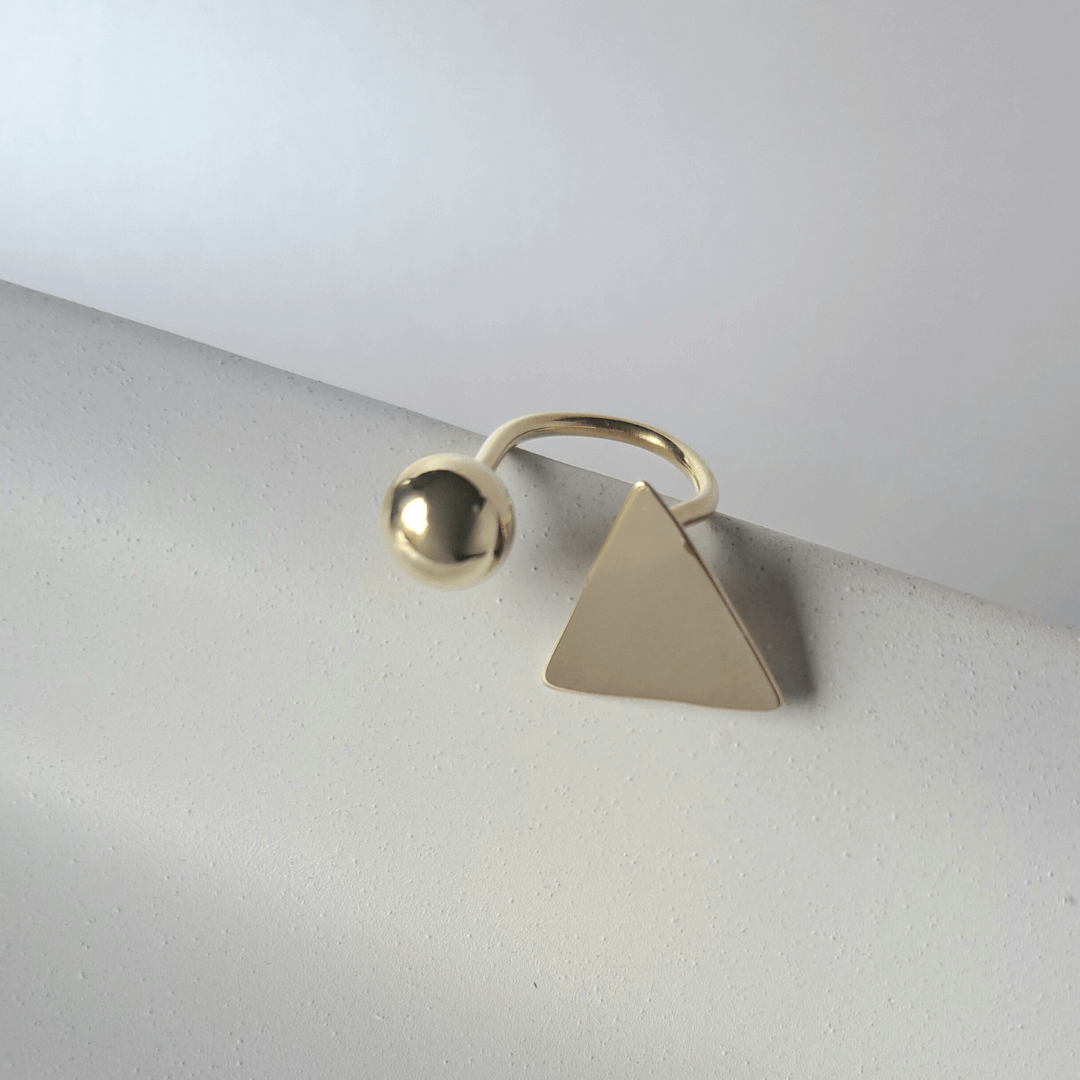 Soline gold ring by Bold Lines Jewellery, featuring a triangle and ball design, displayed on a white geometric stand.