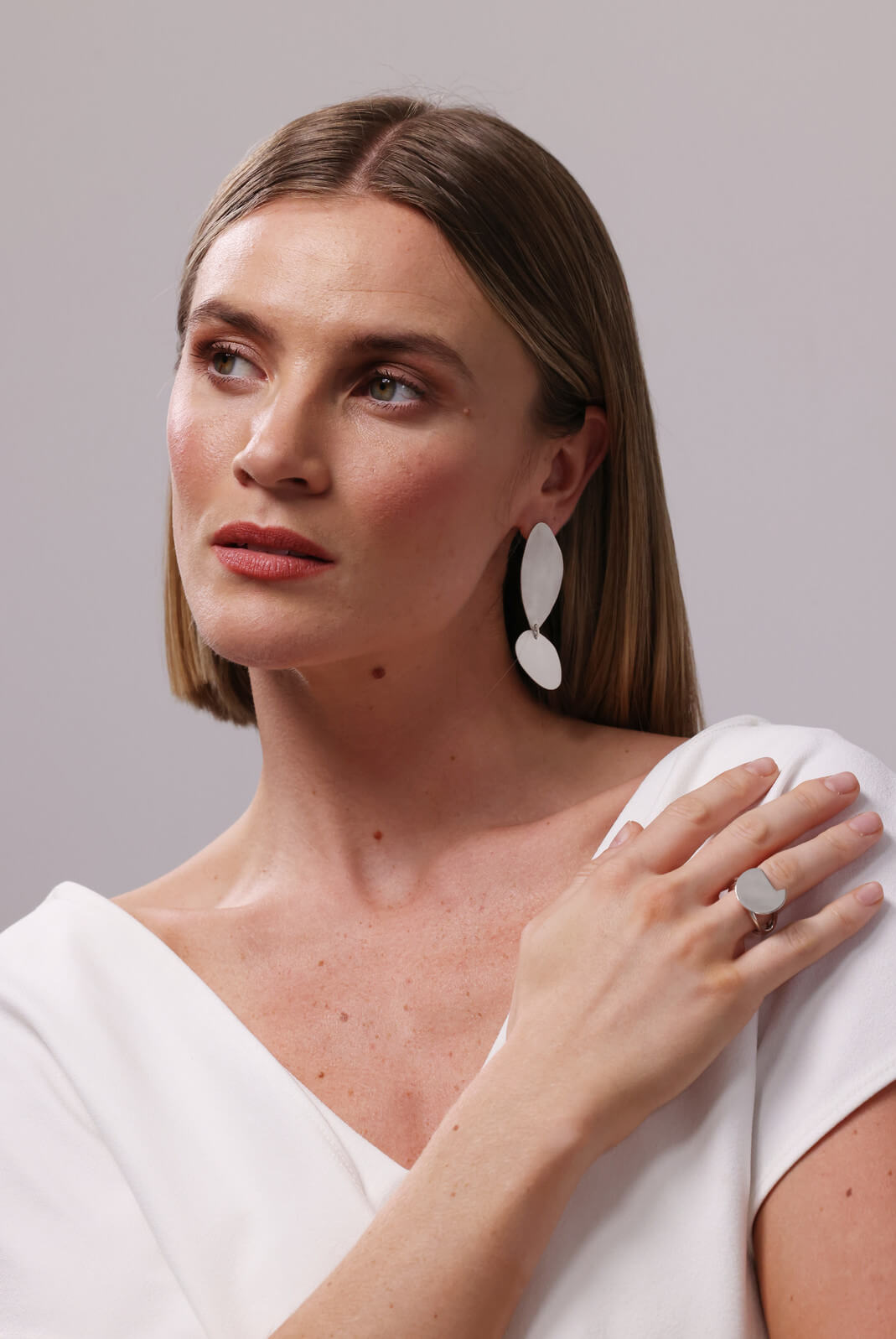 Woman wearing, white, layered  elongated sterling silver earrings and a ring on a neutral background