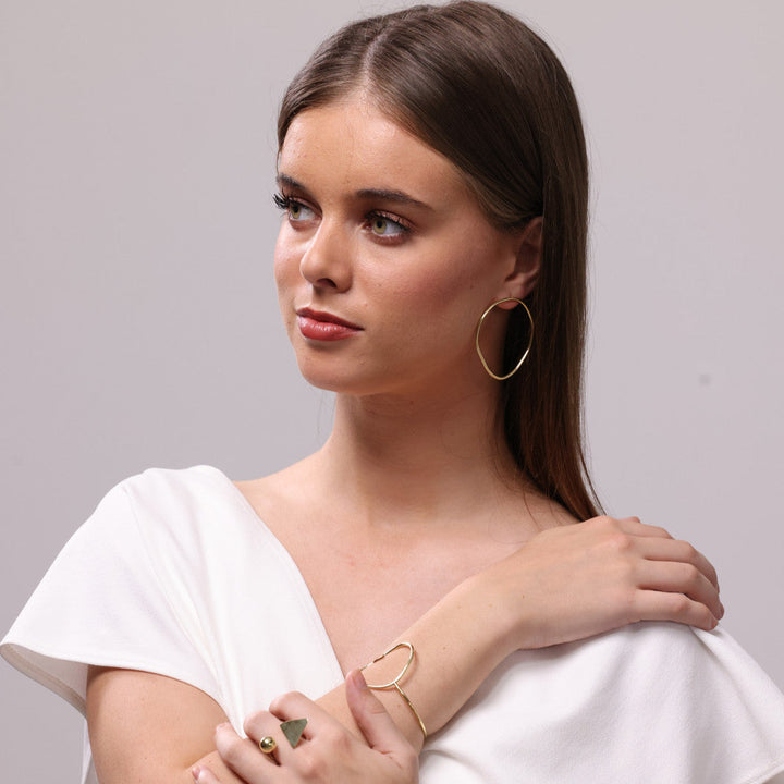 Woman wearing gold wavy hoop earrings, a gold heart-shaped bracelet and a triangle ring on a grey background