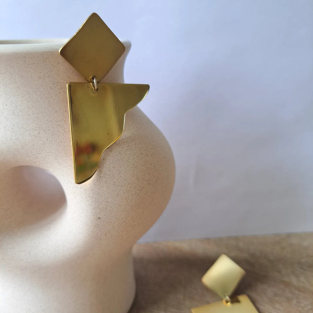 A pair of gold earring, one piece in a ceramic vase the other one is on the table.