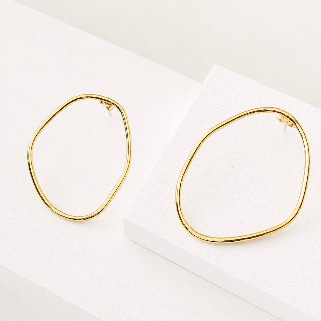 A pair of gold-toned, irregularly shaped wavy hoop earrings displayed on a clean white geometric surface.