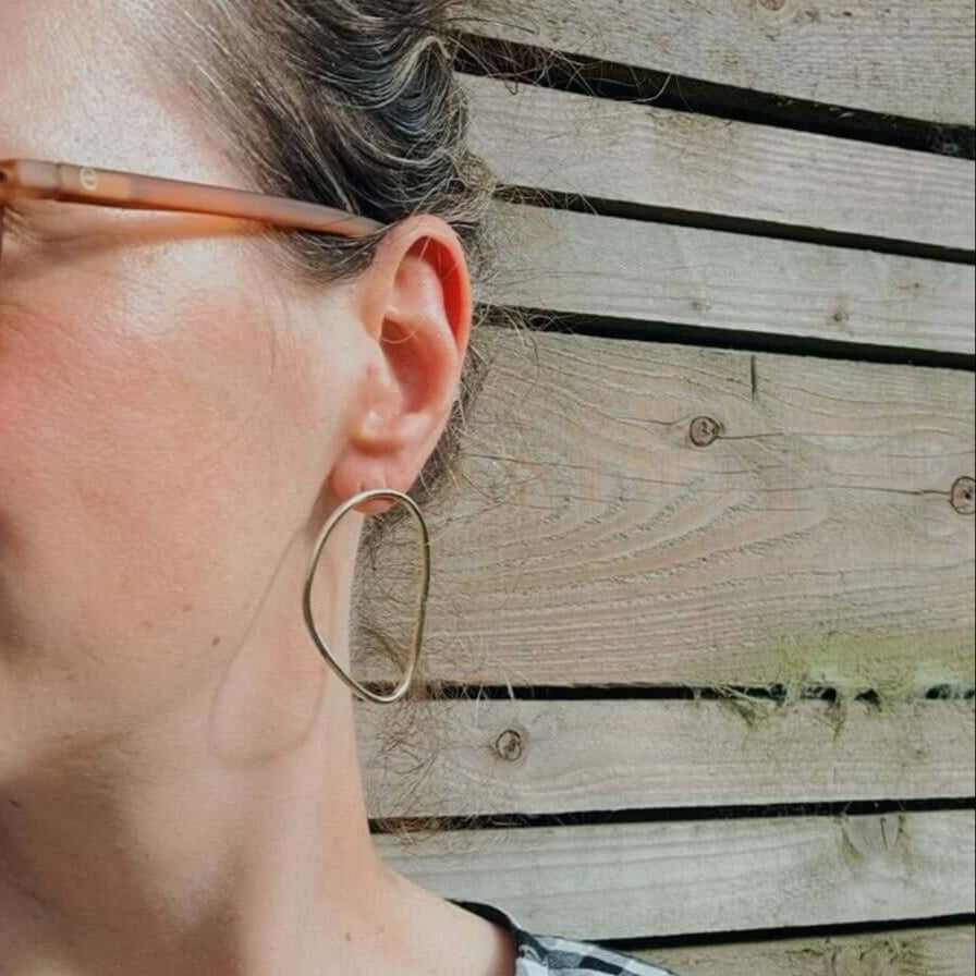 Imperfection organic hoop earrings with sterling silver backs designed by Bold Lines Jewellery worn by a model who is also wearing sunglasses and standing in front of wooden panels.