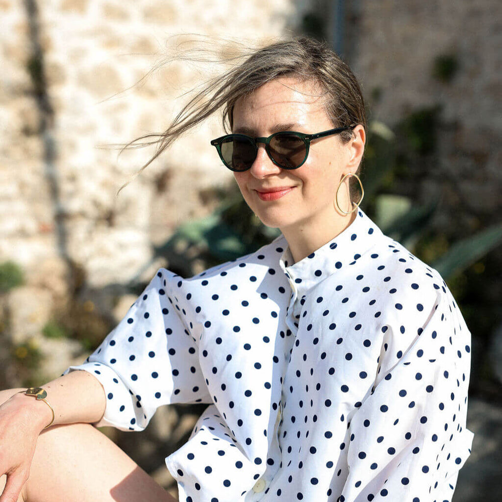Woman wearing a white polka dot shirt the Imperfection gold earrings and green sunglasses outdoors.