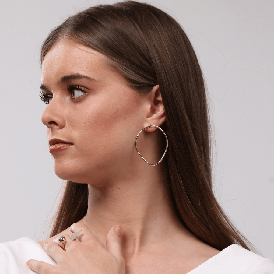 A woman with long brown hair wearing large, thin gold hoop earrings by bold Lines jewellery and a delicate gold bracelet, posing in a white top against a light gray background.