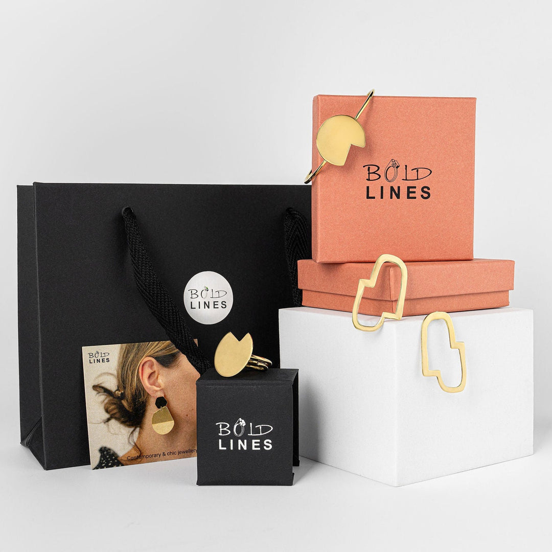 Jewellery packaging with 'Bold Lines' terracotta and black boxes branding on a white background and 3 gold plated jewellery pieces displayed on  white blocks and boxes