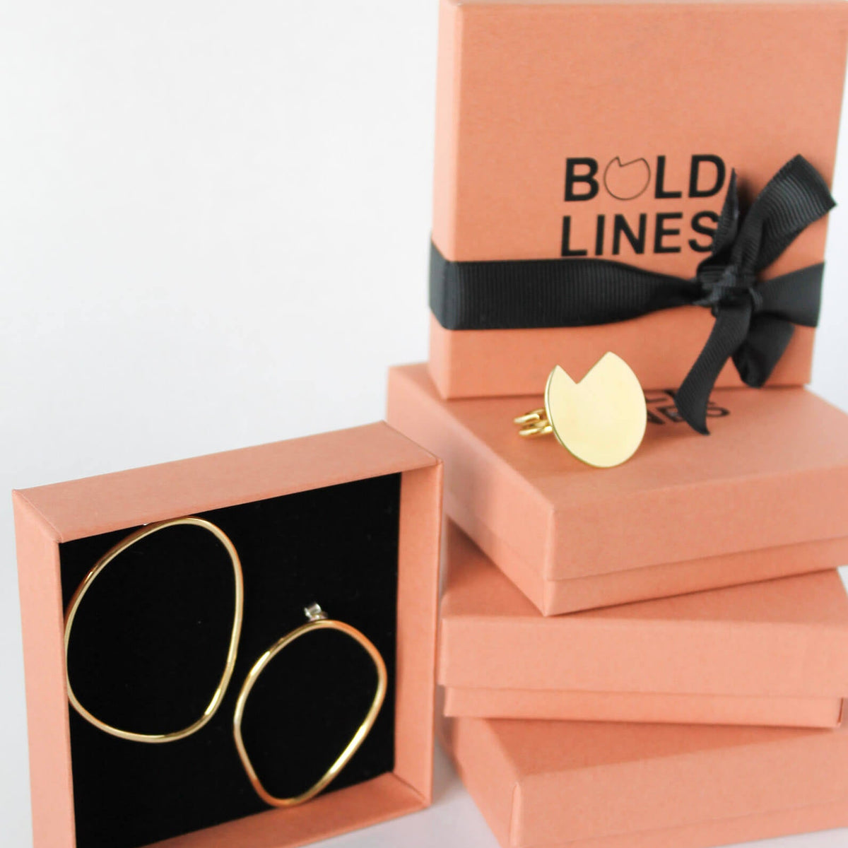 Gold line hoop earrings displayed in an open peach-colored jewelry box next to a stack of matching closed boxes, one of which features a black ribbon and the brand name 'BOLD LINES'.