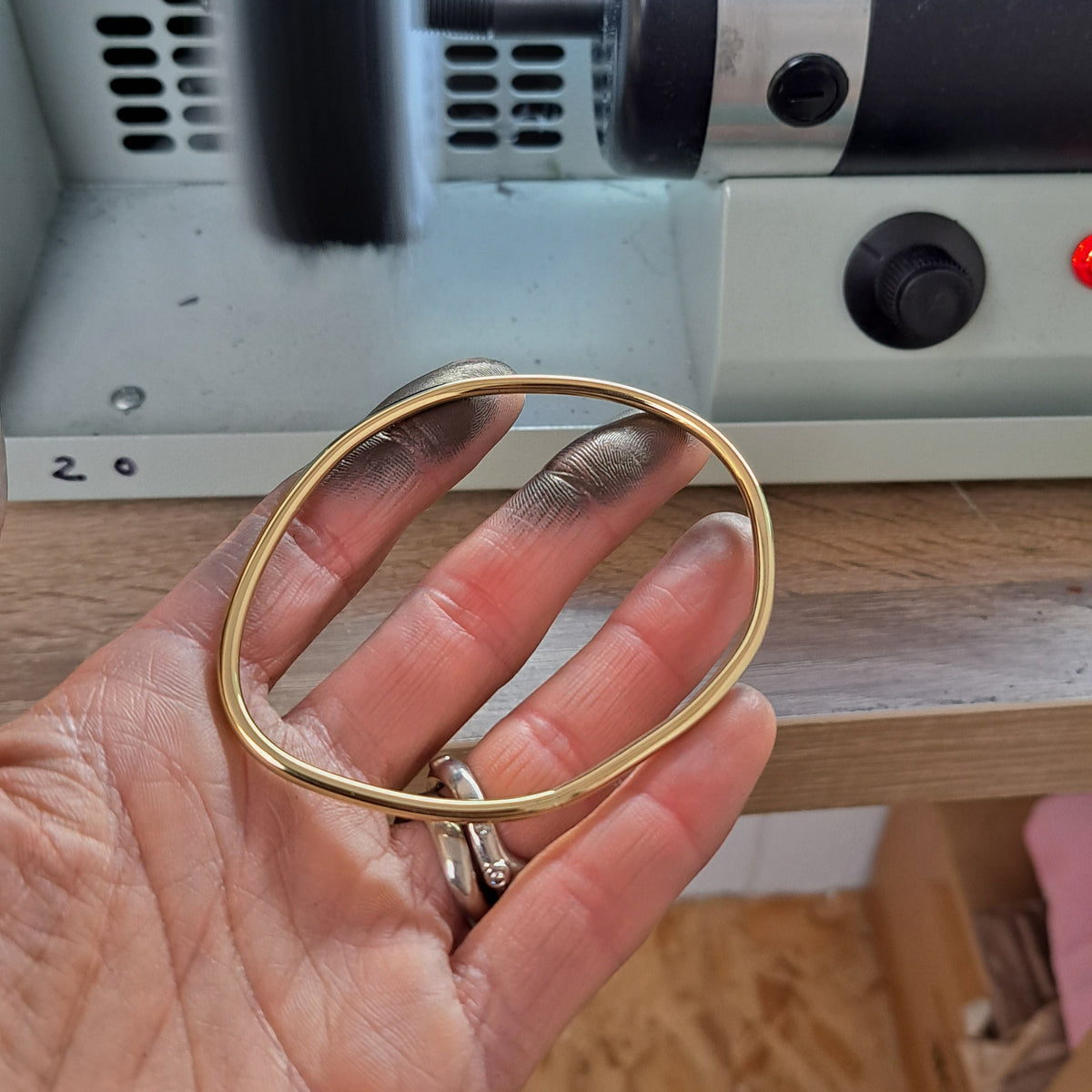 A hand holds a thin gold bangle bangle by bold lines jewellery in front of a small metal rotary sharpening or buffing machine.