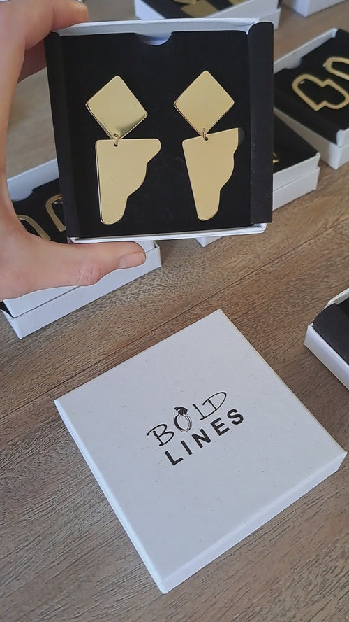 A video of Bold Lines Dream II earrings in a black lined box being held up. The lid of the box lies below.
