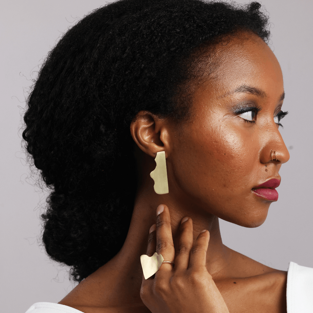 A woman in profile wearing minimalist gold geometric earrings and a matching gold ring, set against a solid light gray background.
