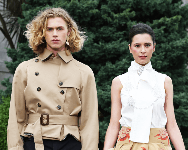 Two models wearing fashion outfits standing outdoors with trees and a building in the background.